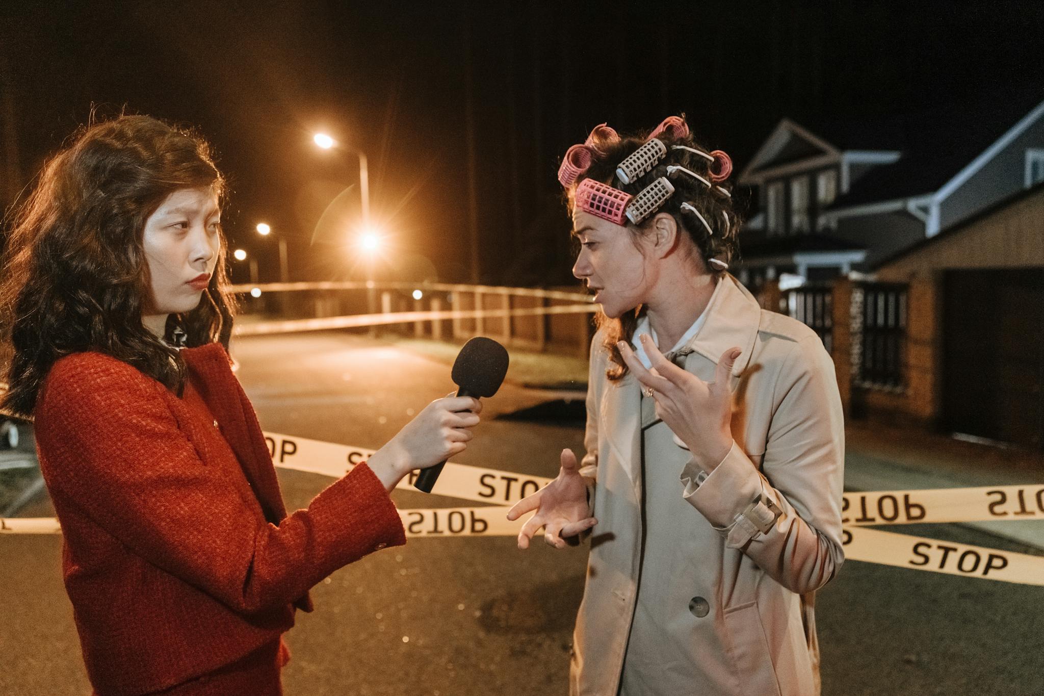 Asian female journalist interviews a woman at a night crime scene. Police tape marks the area.